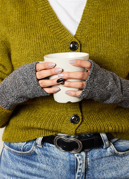 Person wearing gray hand-knitted fingerless gloves, styled with a mustard yellow cardigan and blue jeans, holding a white cup – cozy and functional knitwear for everyday use.