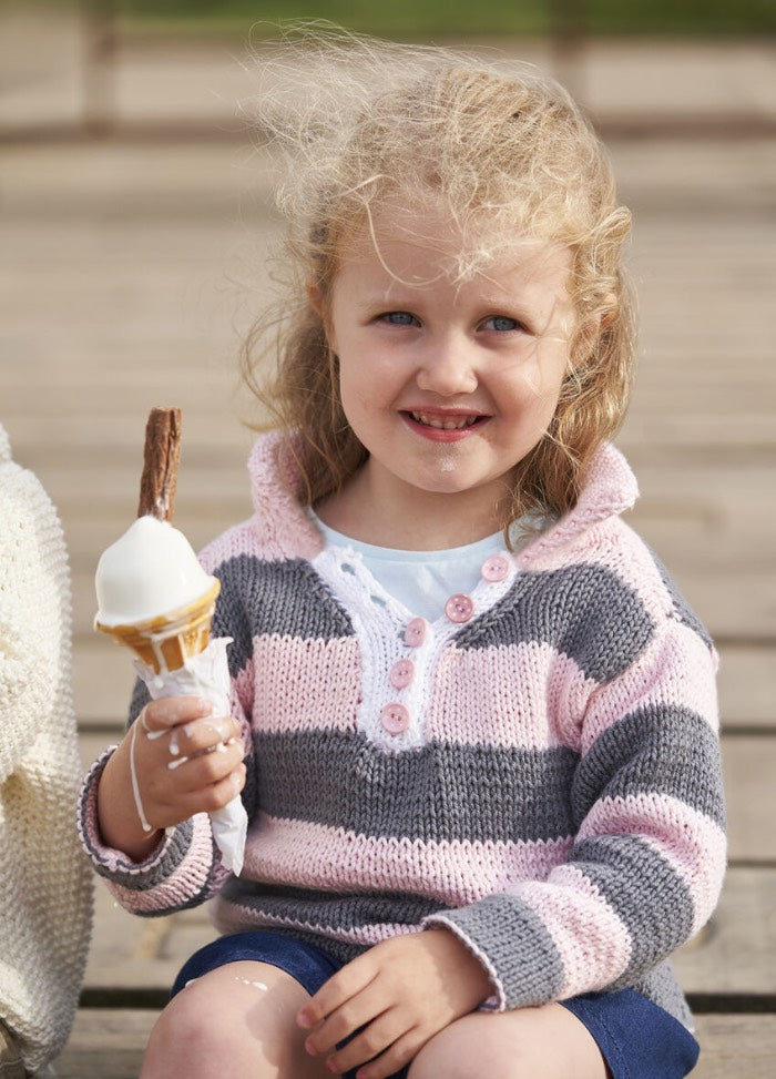 A young girl with curly blonde hair smiles while holding an ice cream cone topped with a chocolate flake. She wears a pink and gray striped sweater.