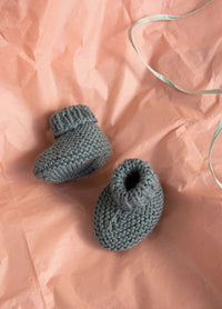 Gray knitted baby booties rest on crumpled peach paper with a silver ribbon. The scene feels soft and cozy, evoking a sense of warmth.