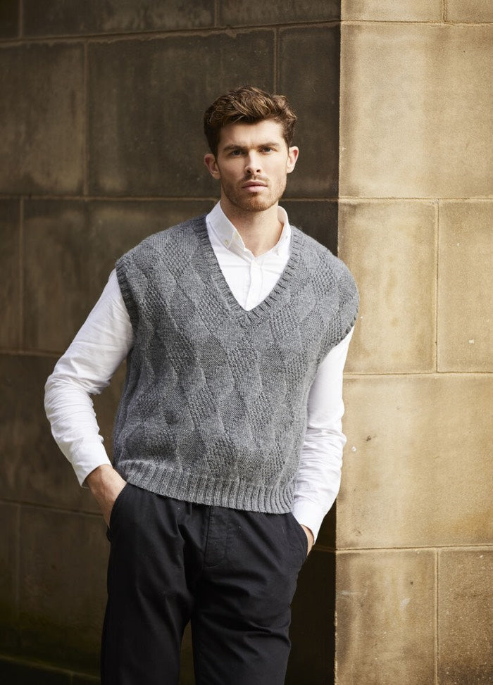 Man in a gray knitted vest over a white shirt leans against a stone wall. His expression is thoughtful, creating a sophisticated and relaxed mood.