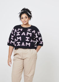 Woman in beige pants and a black sweater with "I AM" text, standing confidently. Her hair is curly, tied with a bow. Neutral background.