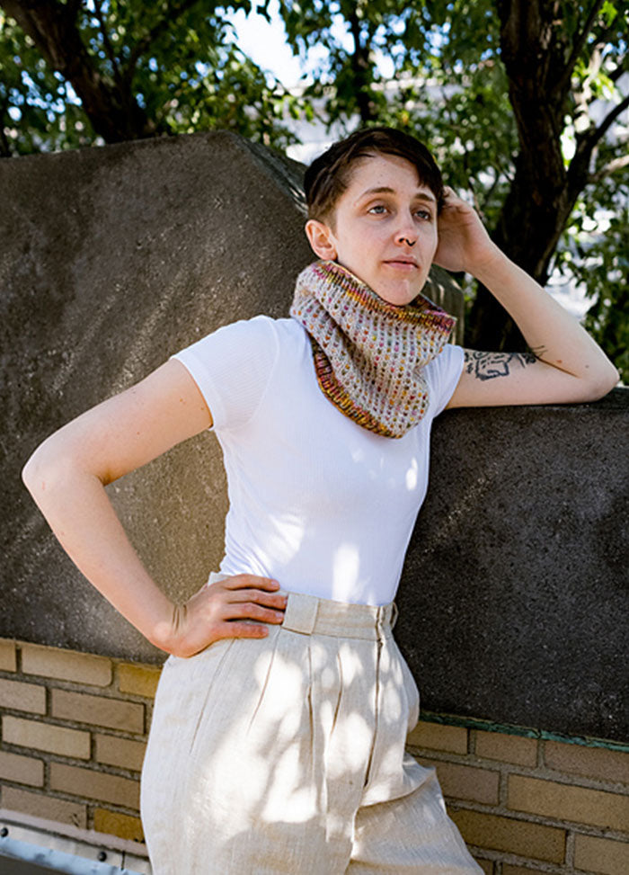 Model wearing a pastel Ice Cream + Sprinkles Cowl with a speckled knit pattern, styled with a white tee and light pants, outdoors by a stone wall.