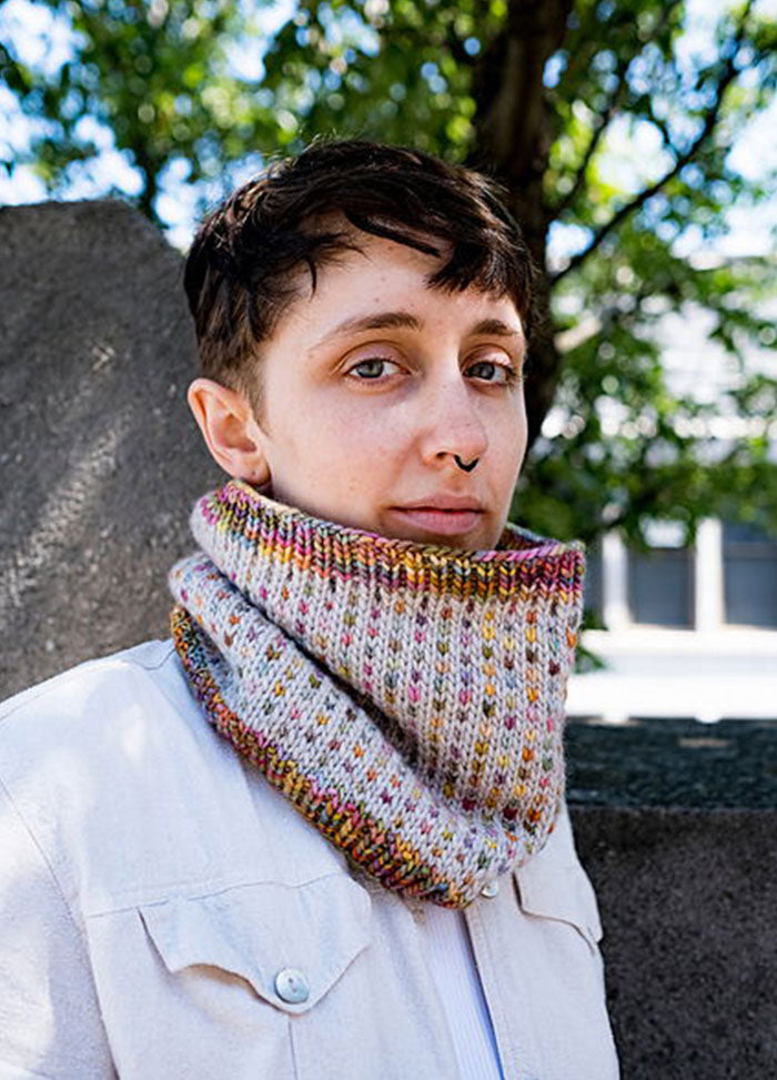 Pastel Ice Cream + Sprinkles Cowl with a speckled knit pattern, worn outdoors with a white tee and light trousers, leaning against a stone wall.