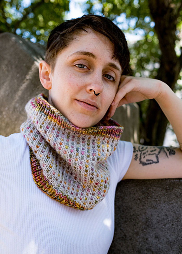 Model wearing a colorful Ice Cream + Sprinkles Cowl with a speckled pastel pattern, photographed outdoors near stone structures and trees.
