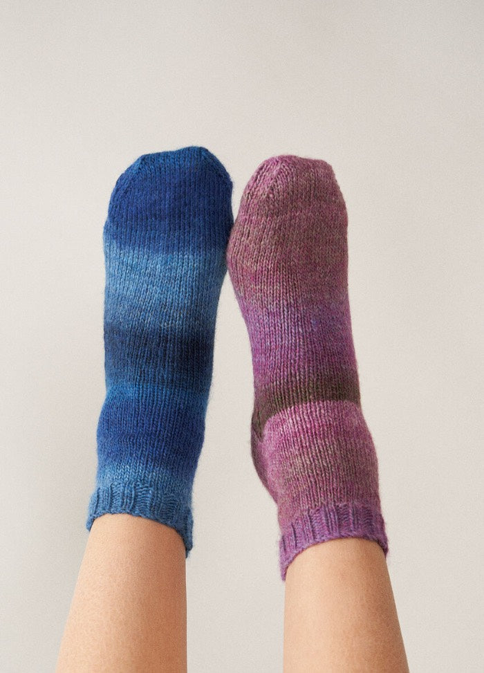 Two feet in mismatched wool socks are raised against a plain background. The left sock is blue, and the right is purple, creating a whimsical, cozy mood.