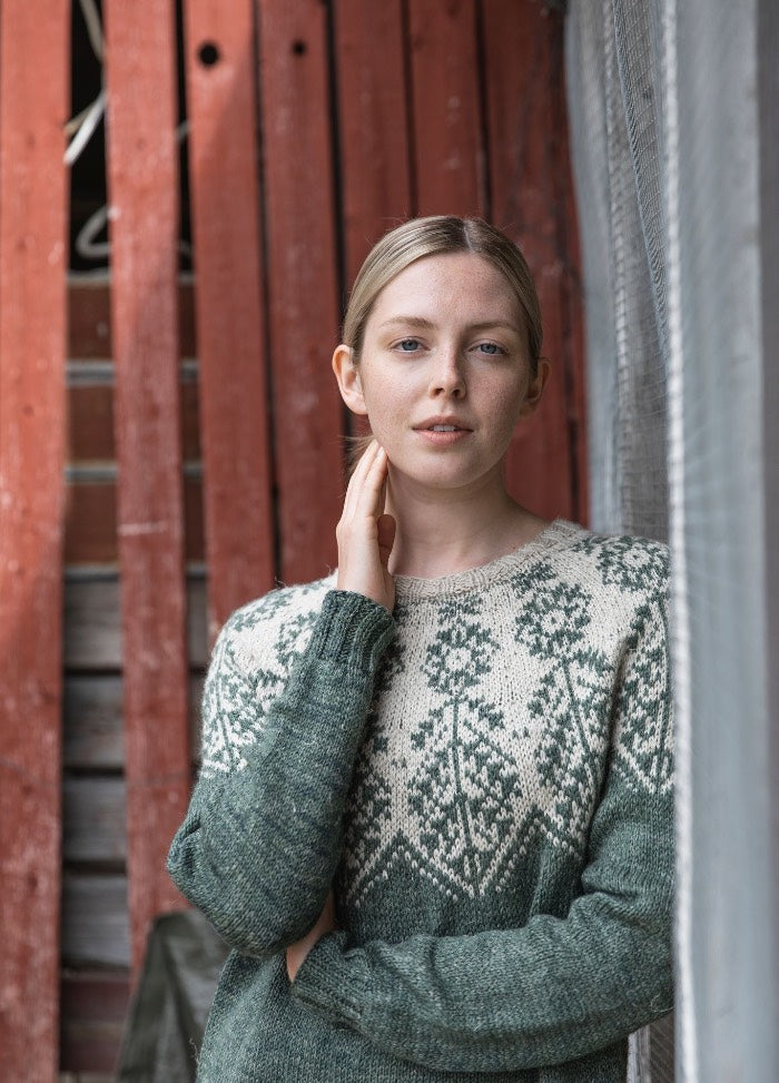 Ilmatar Sweater: green knit with white floral and geometric motifs across the yoke, set against a red wooden wall with metal mesh.