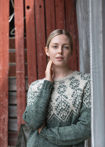 Ilmatar Sweater: green knit with white floral and geometric motifs across the yoke, set against a red wooden wall with metal mesh.