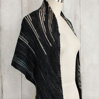 Incremento Shawl draped on a mannequin, featuring dark tones with subtle lighter stripes, shown against a wooden background.