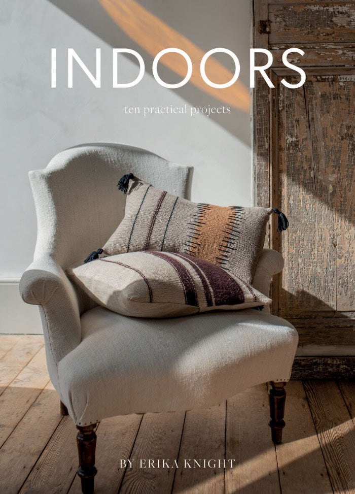A cozy armchair with patterned cushions sits on a wooden floor, bathed in warm sunlight. The background shows a rustic wooden door.
