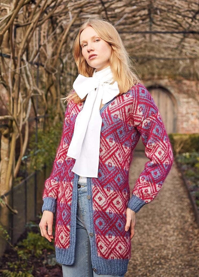 A person in a garden archway wearing a red and blue patterned cardigan over a white blouse with a large bow. The mood is stylish and confident.