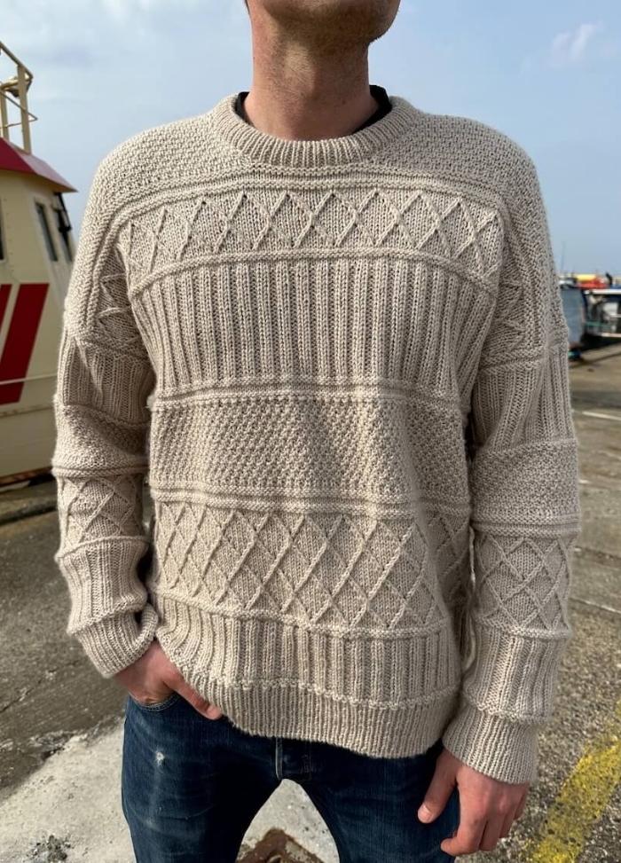 A person wearing a cream-coloured, textured knit sweater with geometric patterns stands outdoors. They have their hands in the pockets of dark jeans.