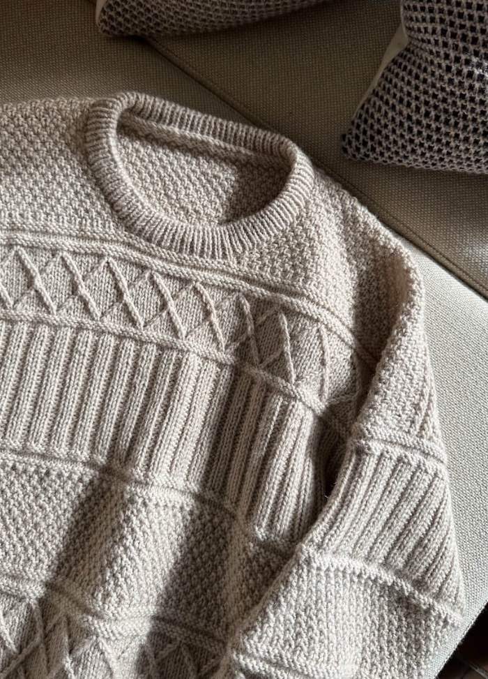 A beige knitted sweater with geometric patterns lies on a light sofa, next to textured grey pillows. Sunlight casts soft shadows, creating a cozy ambiance.