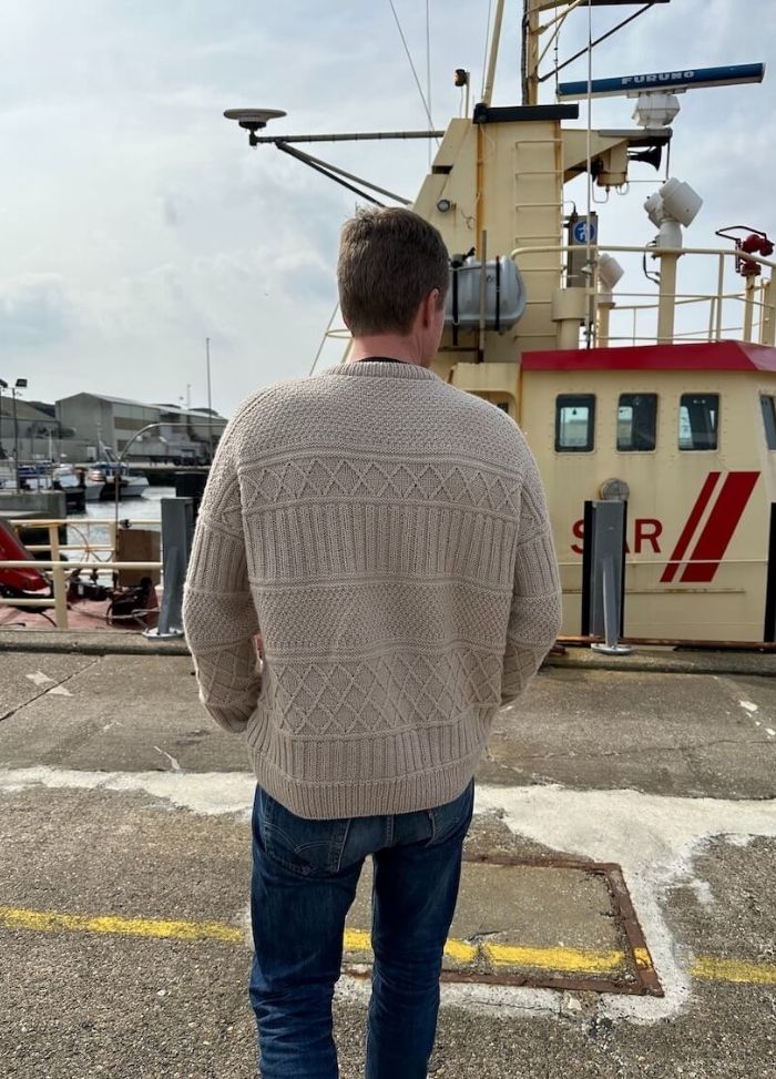 A person in a patterned beige sweater and jeans stands near a yellow and red boat in a harbour. The image conveys a calm, overcast day.