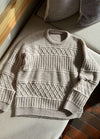 Cozy beige knitted sweater with intricate patterns, including diamond and ribbed designs, lying on a textured beige sofa near patterned cushions.