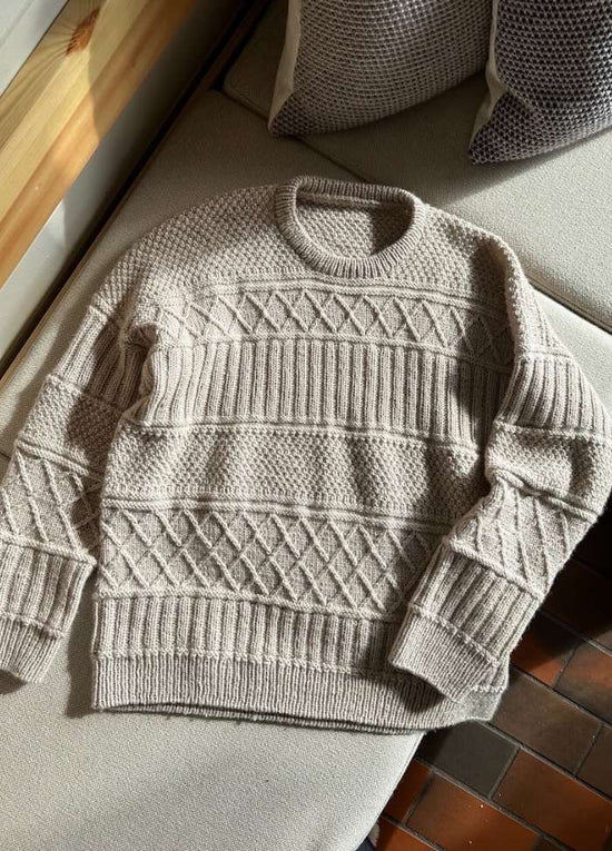 Cozy beige knitted sweater with intricate patterns, including diamond and ribbed designs, lying on a textured beige sofa near patterned cushions.