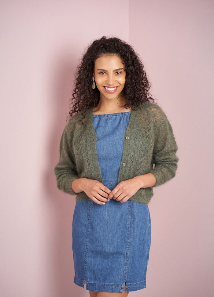 Smiling woman with curly hair in a denim dress and green knit cardigan stands against a soft pink background, conveying warmth and style.