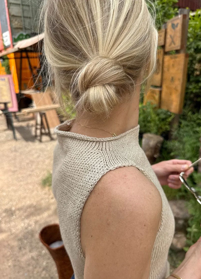 Side view of the Ivy Top in sleeveless beige knit with high neckline, worn outdoors with wooden signs and greenery in the background.