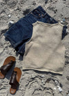 Flat lay of the Ivy Top in beige knit, paired with dark denim shorts and brown leather sandals on a sandy beach background.