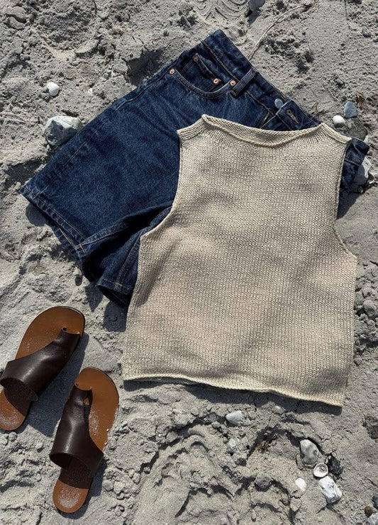 Flat lay of the Ivy Top in beige knit, paired with dark denim shorts and brown leather sandals on a sandy beach background.