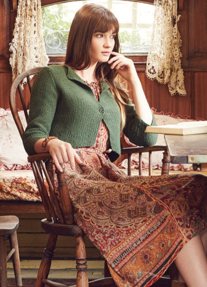 Model wearing the Jampa Jacket from the Rowan Softyak DK Collection by Marie Wallin, a green knitted jacket layered over a patterned dress with intricate designs, photographed indoors in a rustic room with lace curtains, wooden walls, and a window overlooking greenery.