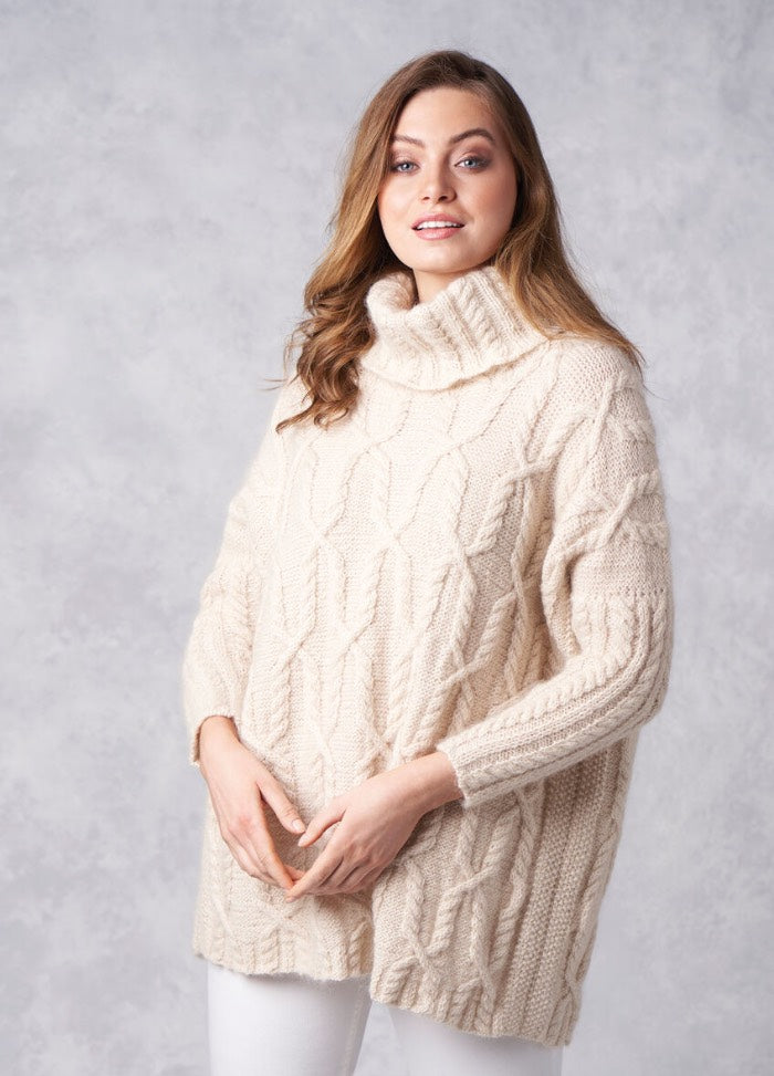 A woman in a cozy, cream cable-knit turtleneck sweater poses against a light gray background. She has wavy brown hair and a gentle, confident smile.