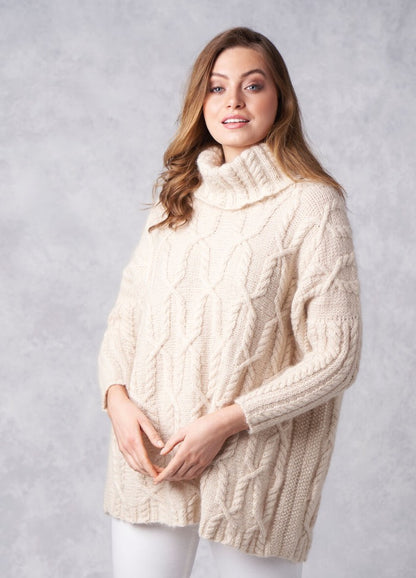 A woman in a cozy, cream cable-knit turtleneck sweater poses against a light gray background. She has wavy brown hair and a gentle, confident smile.