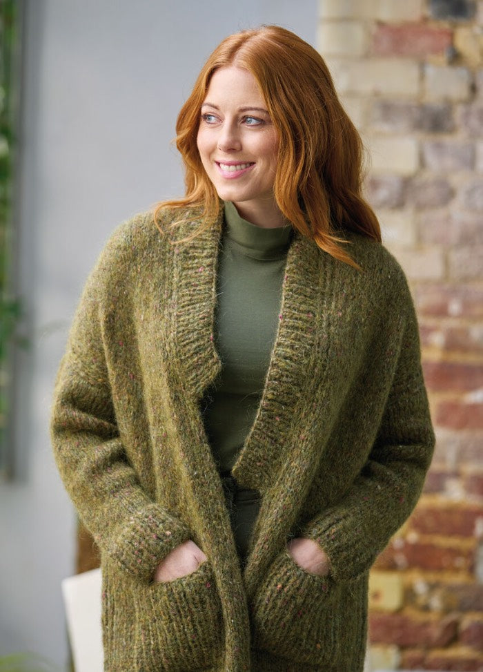 A person with red hair smiles warmly while wearing a thick, green knit cardigan. They have their hands in the cardigan's pockets, with a brick wall in the background.