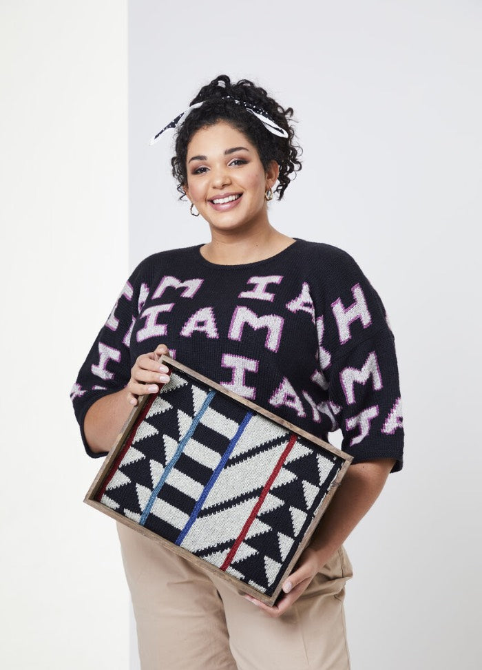 Smiling woman with curly hair wearing a black sweater with "I AM" text, holds a framed geometric art piece. The tone is cheerful and expressive.