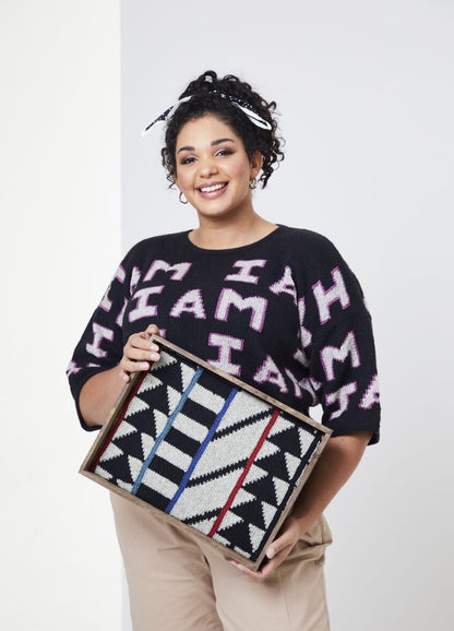 Smiling woman with curly hair wearing a black sweater with "I AM" text, holds a framed geometric art piece. The tone is cheerful and expressive.