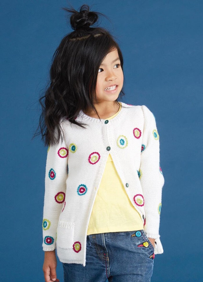 A young girl with long black hair in an updo, wearing a white cardigan with colorful circles, a yellow top, and jeans, smiles slightly against a blue background.