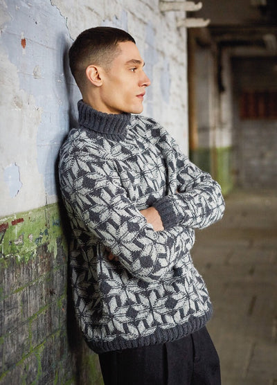 A man in a patterned gray turtleneck sweater and black pants leans against a weathered, painted wall in an industrial setting, appearing thoughtful.