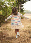 A young girl in a colorful sweater and fluffy skirt walks away down a forest path. Her arms are outstretched, conveying a sense of freedom and joy.
