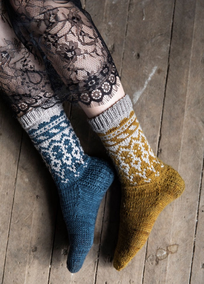 Joukahainen Socks: mismatched blue and mustard yellow socks with white motifs, styled with black lace tights on a wooden floor.