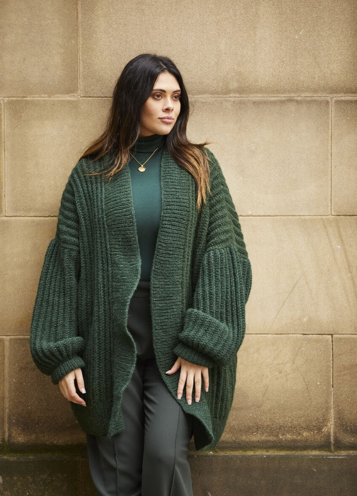 Woman in a cozy, oversized green cardigan and turtleneck stands against a stone wall. Her expression is calm, and she wears a gold necklace.