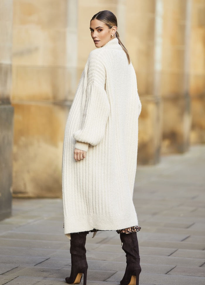 Woman in a long cream knit cardigan and black heeled boots walks on a stone-paved street with a confident gaze over her shoulder. Urban, stylish.