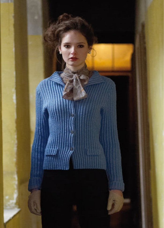 A woman stands in a dimly lit hallway, wearing a blue cardigan, patterned scarf, and gloves, projecting a vintage, contemplative mood.