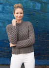 Smiling woman with a bun wearing a gray cable-knit sweater and white pants. She stands against a textured blue background, exuding a casual, cozy vibe.