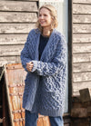 Woman smiling outside, wearing a thick, blue cable-knit cardigan and jeans. She stands against a rustic wooden wall, exuding warmth and coziness.
