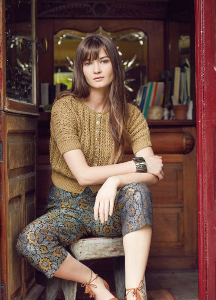 Model wearing the Kaya Cardigan from the Rowan Softyak DK Collection by Marie Wallin, a short-sleeved light brown knit cardigan with front buttons, paired with floral patterned pants, photographed indoors near an open door with wooden furniture and bookshelves in the background.
