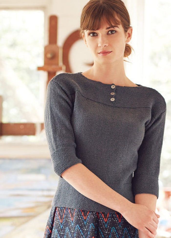Model wearing the Ketu Sweater from the Rowan Softyak DK Collection by Marie Wallin, a grey three-quarter sleeve knit with three buttons at the chest, paired with a zigzag-patterned skirt in blue, red, and black, photographed indoors with a blurred wooden structure in the background.