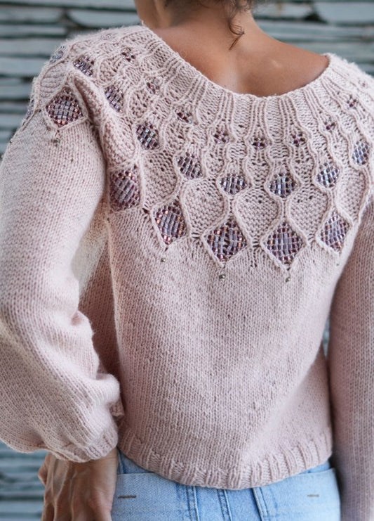 Woman wearing a soft pink knitted sweater with intricate lace patterns and bead details on the back. Light blue jeans complete the casual, stylish look.