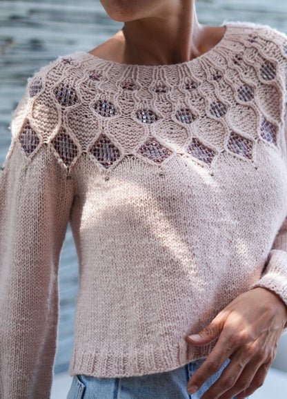 Woman wearing a light pink sweater with intricate lacework and sequin details on the yoke, paired with faded blue jeans. Soft and cozy vibe.