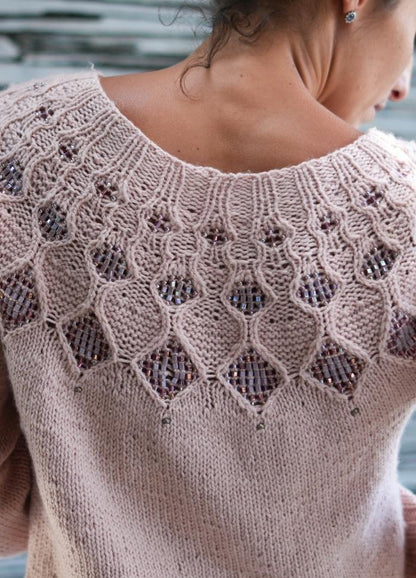 A woman wears a soft pink knitted sweater featuring intricate lace patterns with embedded beads. The design adds an elegant, delicate touch.