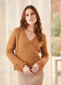 Woman with long brown hair wearing a warm brown sweater with lace cuffs and a beige skirt. She stands in a softly lit room with a calm expression.