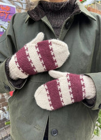 A person wearing a green coat and brown sweater shows off cozy knit mittens with maroon and cream stripes, conveying a warm, rustic vibe.