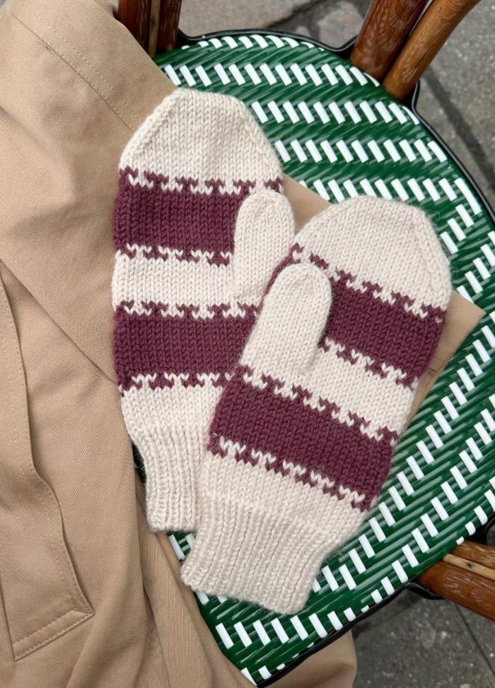 Cozy knit mittens with burgundy and cream stripes, lie on a tan jacket draped over a woven green and white chair.