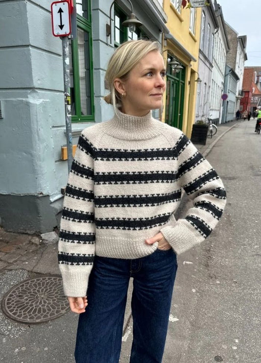 A woman stands on a quaint street wearing a beige sweater with black stripes and dark jeans.