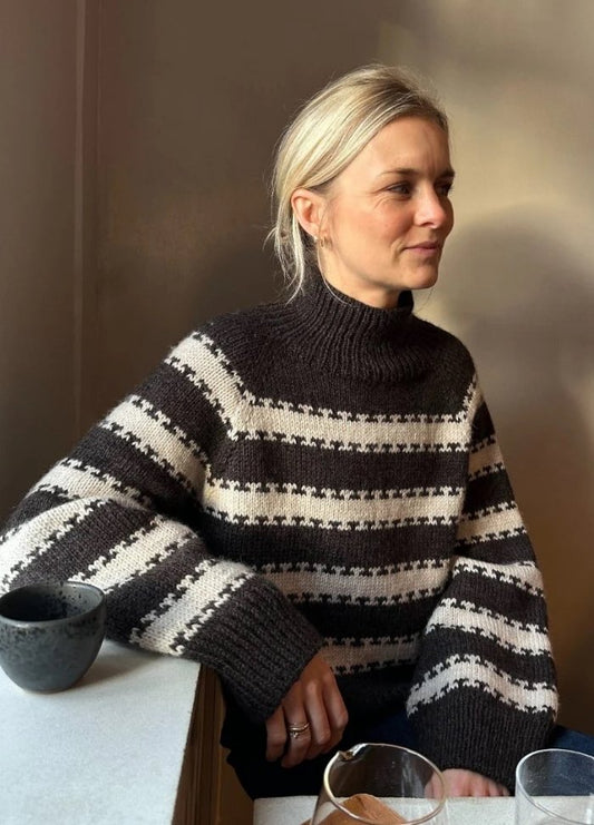 A woman with blonde hair sits at a table, wearing a black and white striped sweater. Soft, warm lighting creates a relaxed atmosphere.
