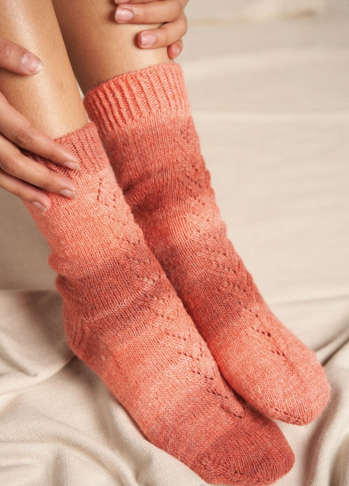 Close-up of cozy, hand-knit orange socks with a textured pattern worn by a person sitting cross-legged on a soft, cream-colored blanket.