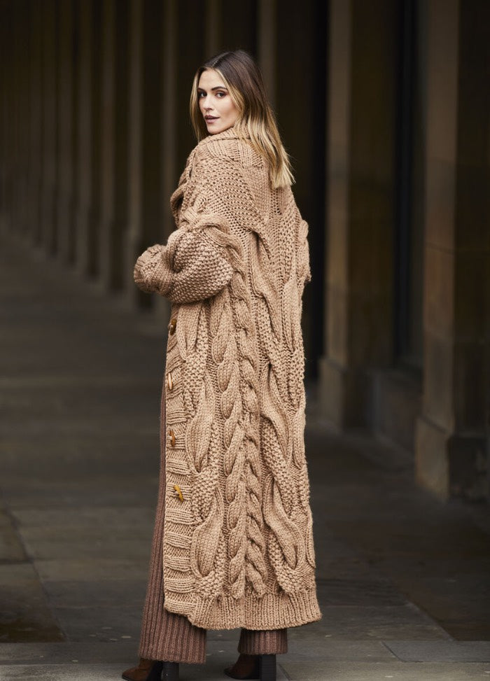A woman in a long, chunky, beige knitted cardigan with intricate cable patterns walks down a dimly-lit, column-lined corridor, exuding a cozy, stylish vibe.